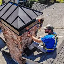 Two workers in safety harnesses repairing a red brick chimney affected by efflorescence on chimney bricks, with a black metal cap, situated on a shingled rooftop under bright daylight.