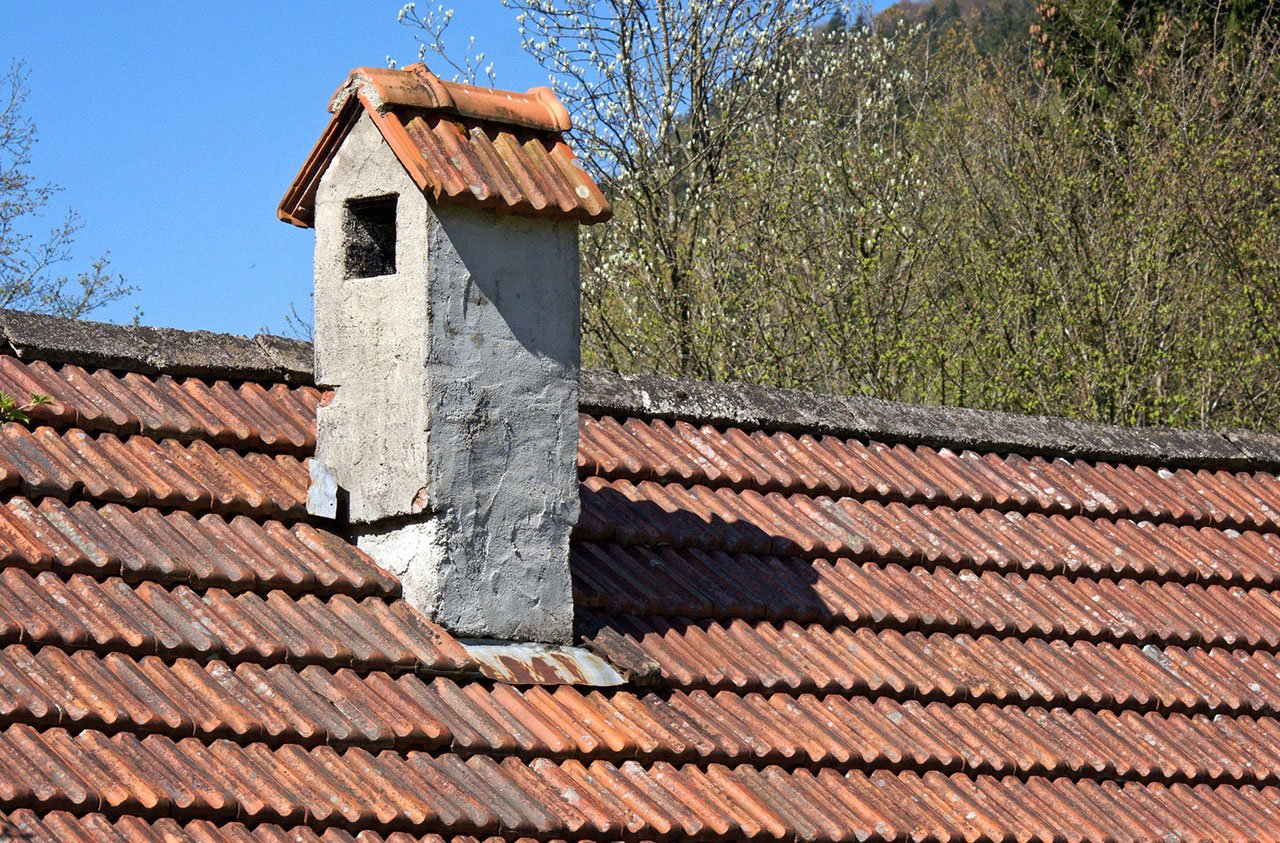 A close-up view of a brick chimney with visible signs of wear, including cracks and peeling mortar. Moss and ivy are growing along the chimney, indicating long-term exposure to moisture. The surrounding area includes part of a roof and cloudy sky in the background, emphasizing the weathered condition.