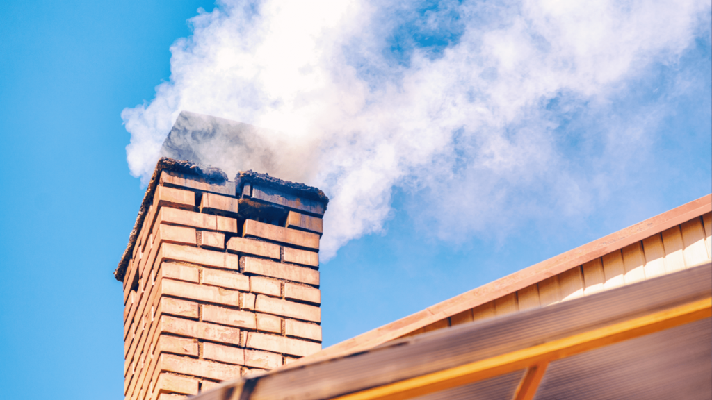 A close-up view of a chimney with visible rust and weathering, showing signs of deterioration and wear. The chimney cap appears worn, with corrosion marks, and there is visible discoloration along the pipe. The sky in the background is overcast, emphasizing the structure's age and exposure to the elements.