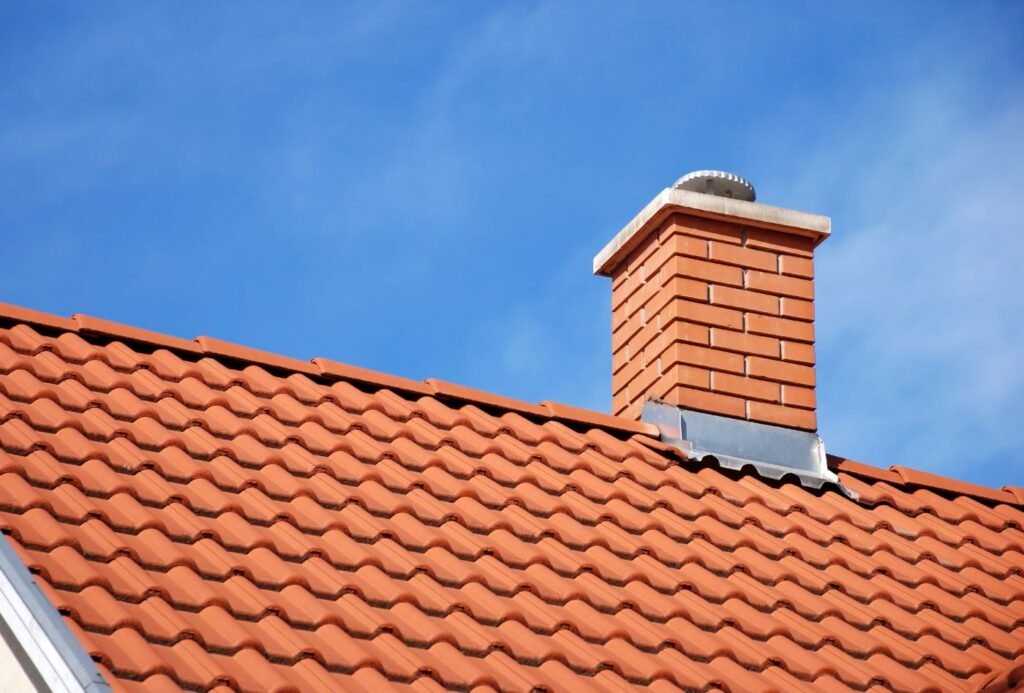 A brick chimney with a metal flue cap is partially visible, showing soot buildup and signs of weathering. The surrounding bricks are slightly eroded, with the sky in the background creating a soft contrast against the chimney's texture.