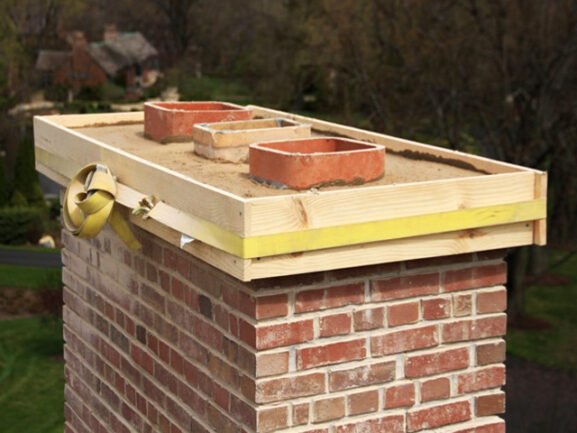 A brick chimney with a wooden formwork secured by a yellow strap, ready for a concrete crown pour, with three clay flue liners visible.
