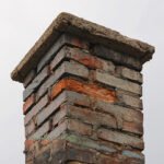 A close-up of a weathered brick chimney with crumbling mortar, some exposed orange brick, and a damaged concrete cap against a grey sky.