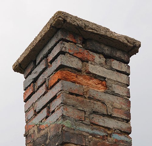 A close-up of a weathered brick chimney with crumbling mortar, some exposed orange brick, and a damaged concrete cap against a grey sky.