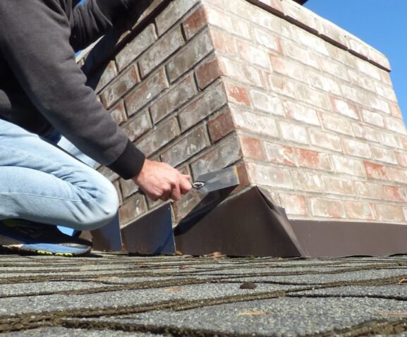 A person in jeans and a dark shirt uses a trowel to apply sealant or mortar around a brown metal flashing at the base of a brick chimney on a shingled roof.