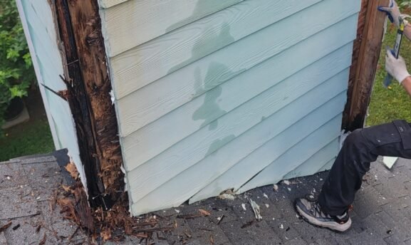 A damaged chimney chase siding with light blue siding peeling away to reveal rotted wood framing and debris on the roof.