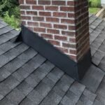 A red brick chimney with new black metal step flashing and counter flashing installed on a grey shingle roof.