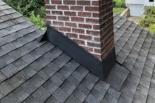 A red brick chimney with new black metal step flashing and counter flashing installed on a grey shingle roof.