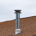 A tall, grey metal chimney flue pipe with a rain cap extending from a brown shingle roof under an overcast sky.