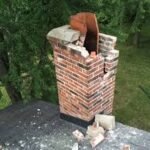 A severely damaged brick chimney with a large vertical crack, broken bricks, and a partially collapsed flue liner, standing on a dark shingle roof amidst green trees.