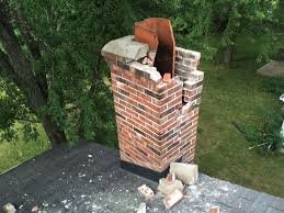 A severely damaged brick chimney with a large vertical crack, broken bricks, and a partially collapsed flue liner, standing on a dark shingle roof amidst green trees.